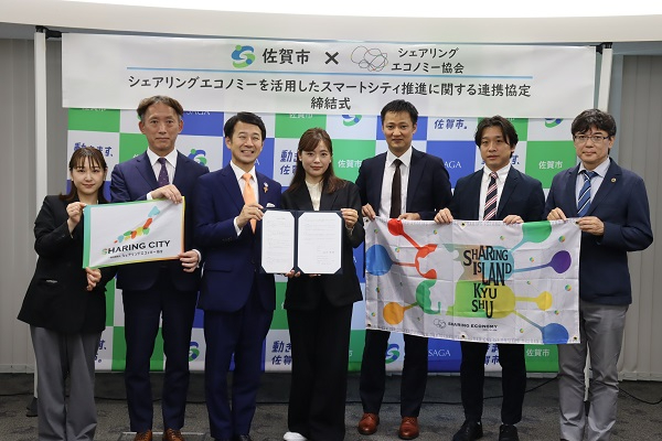 佐賀市とシェアリングエコノミー協会の連携協定締結式にて、横断幕を背にスーツ姿の男女6名が並び、中央の二人が協定書を、左右の人々が「SHARING CITY」などのロゴ入りフラッグを掲げている集合写真