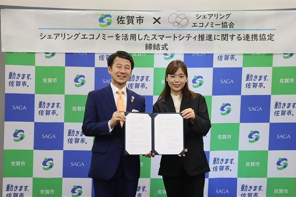 佐賀市とシェアリングエコノミー協会のロゴが並んだバナーを背に、スーツ姿の市長と代表の女性が「シェアリングエコノミーを活用したスマートシティ推進に関する連携協定締結式」と書かれた協定書を二人で持つ写真