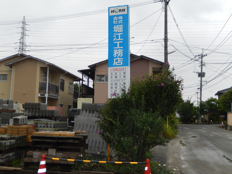 道路沿いに設置された縦長の大きな看板に「株式会社 堀江工務店」と青地に白抜きの大きな文字で社名が記され、その下には左官工事や外構工事などの営業品目が並び、周囲にはコンクリートブロックなどの資材が積まれた作業場の写真