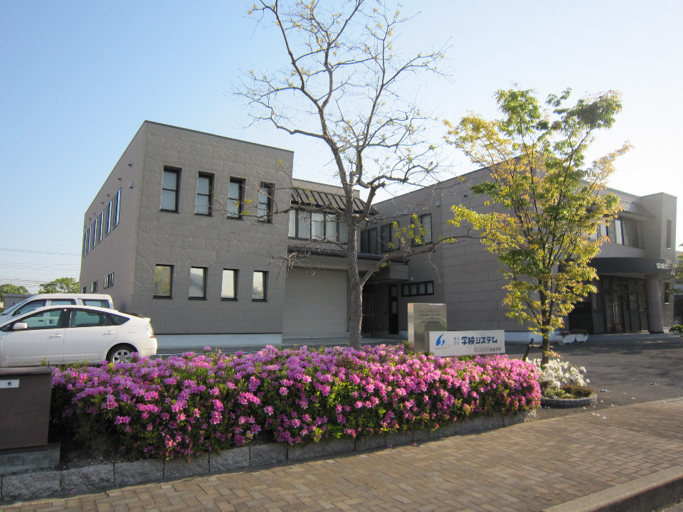 手前に鮮やかなピンク色のツツジが咲き誇る花壇があり、奥には「株式会社学映システム」の看板とグレーの外壁の二階建てのオフィスビルが建ち、白い乗用車が駐車場に停まっている外観写真