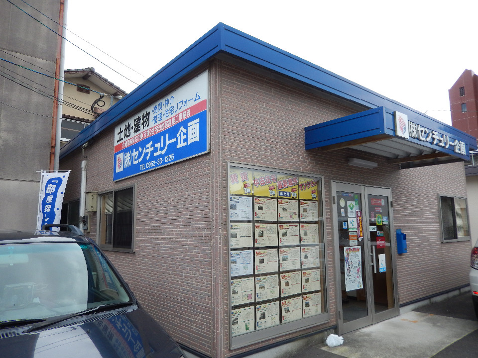 茶色のレンガ調タイルが貼られた平屋の建物で、壁面に「土地・建物」「株式会社センチュリー企画」と書かれた大きな看板を掲げ、窓一面に多数の物件案内チラシが貼り出された店舗外観を捉えた写真