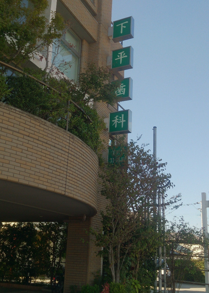 茶色のタイル貼りのビル壁面に、上から一文字ずつ「下」「平」「歯」「科」と書かれた緑色の四角い袖看板が縦に並んで設置されてた下平歯科医院の外観写真