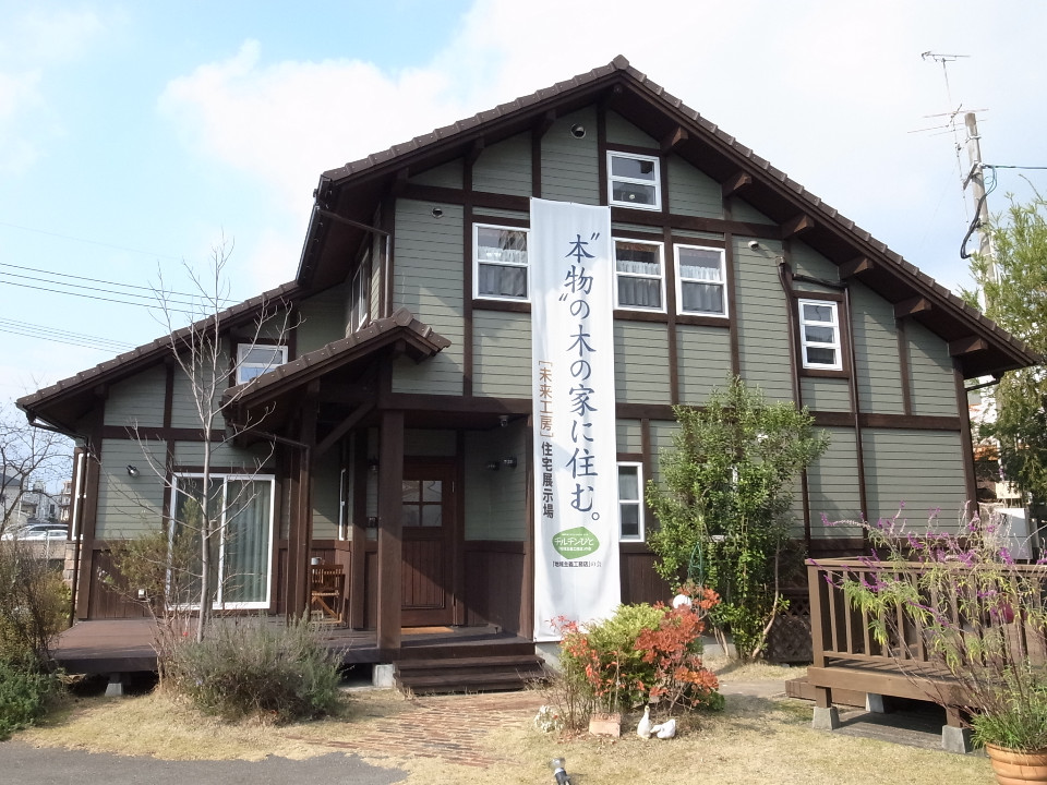 薄緑色の外壁に濃い茶色の柱が映える落ち着いた色調の木造二階建て住宅で、正面には「本物の木の家に住む。」と書かれた大きな白い垂れ幕が掲げられ、植栽やウッドデッキが整えられたモデルハウスの外観写真