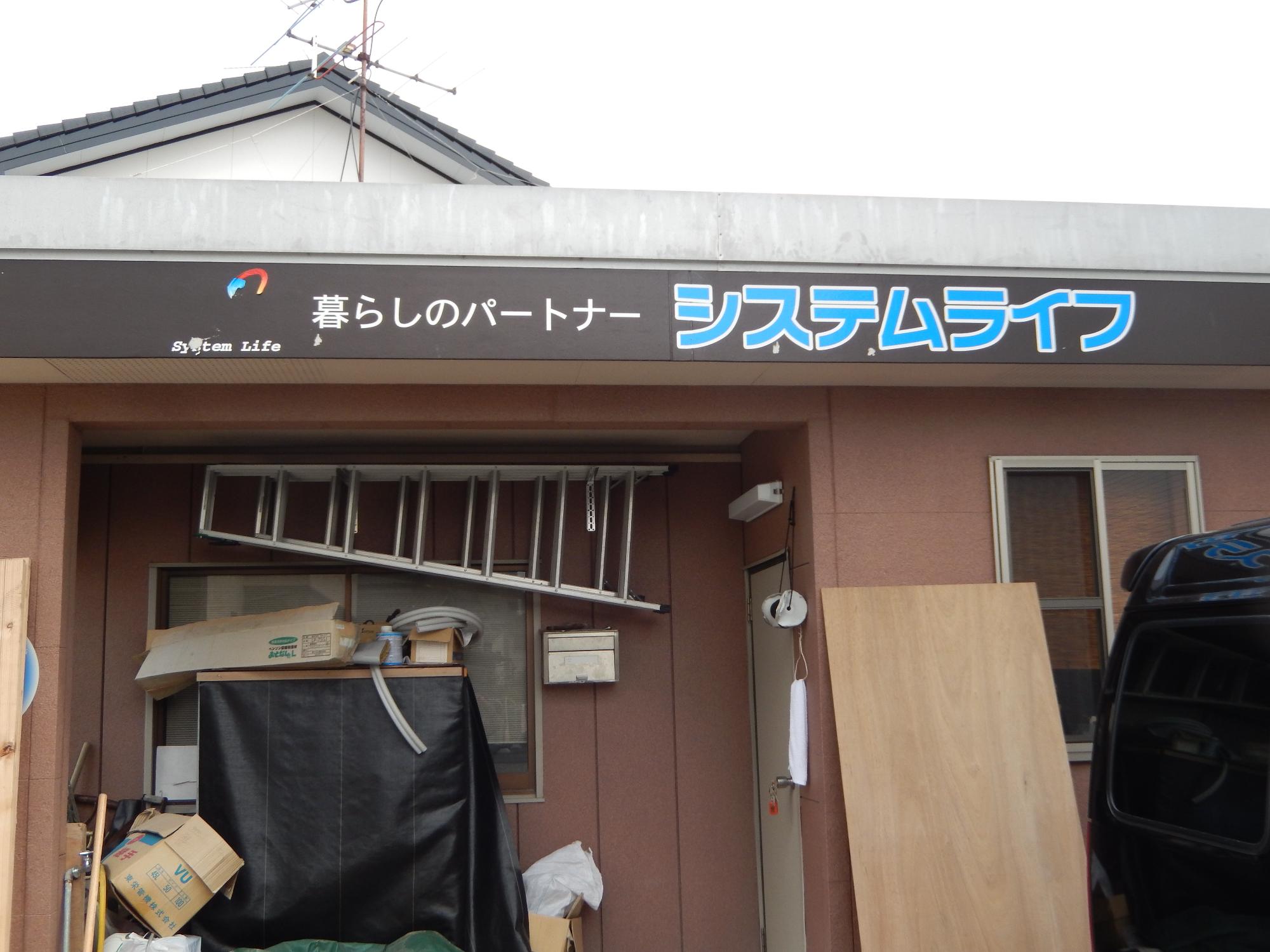 茶色の外壁の建物入口上部に、「暮らしのパートナー システムライフ」というロゴ入りの看板が設置されており、軒下には梯子や作業道具、資材などが置かれている、株式会社システムライフの外観写真