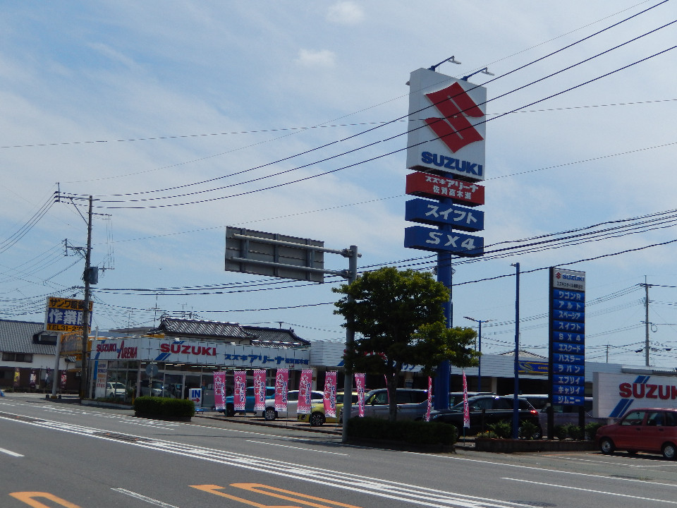 道路沿いに建つスズキスタイル株式会社で、赤い「S」のロゴが大きく描かれた高い看板が立ち、店頭にはピンク色の大売り出しののぼりが多数はためいている活気ある店舗の外観写真