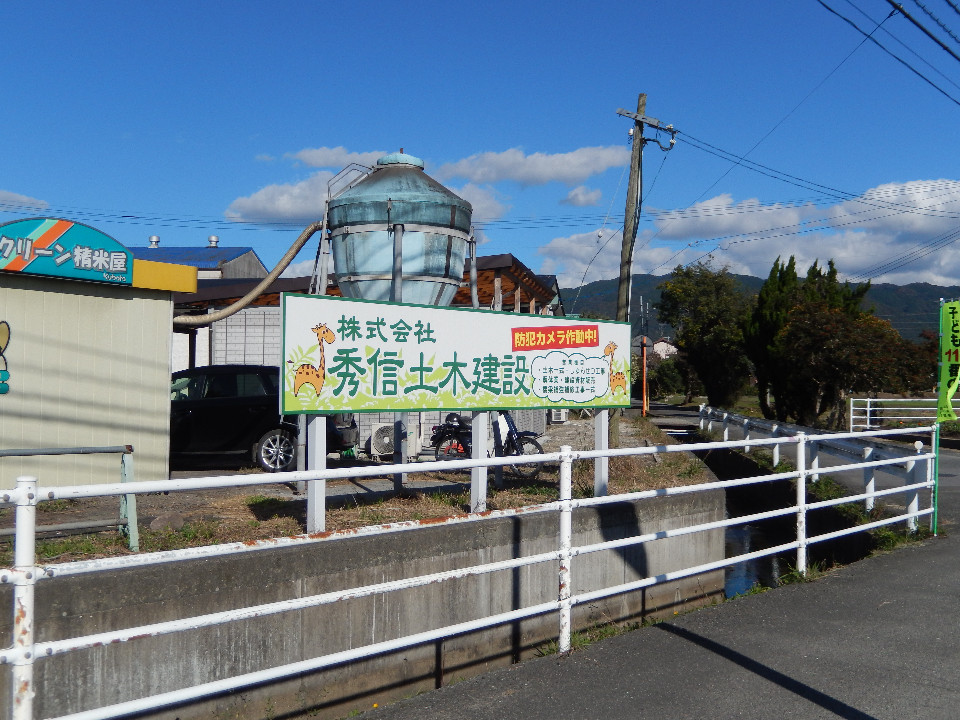 青空の下、白いガードレール越しに株式会社秀信土木建設の看板とコンビニ看板、円形貯水タンクが立ち並んでいる写真
