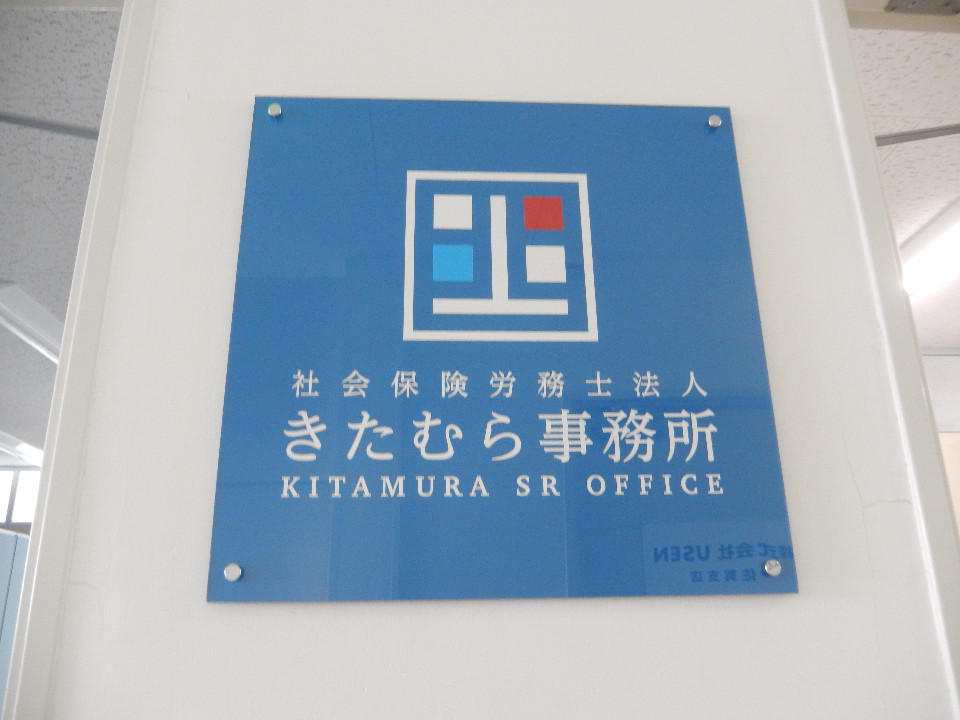 白壁に設置された正方形の青いアクリル板に、白・赤・水色の四角形を組み合わせたシンボルマークと「社会保険労務士法人 きたむら事務所」および英語表記の名称が白色で印字されている看板写真