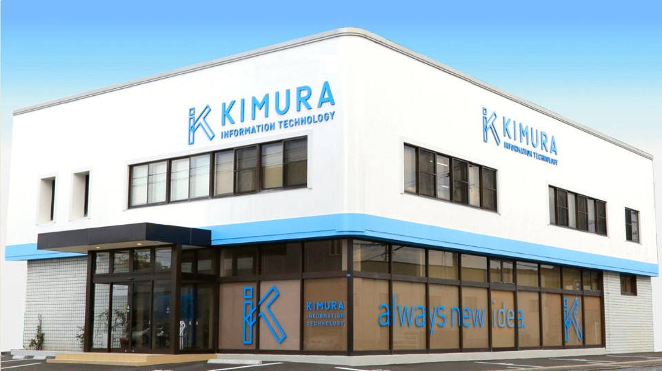 白い外壁と水色のラインが特徴的な二階建てのオフィスビルが建っており、壁面には「KIMURA INFORMATION TECHNOLOGY」の青いロゴが掲げられ、1階の大きなガラス窓にもロゴやメッセージがデザインされている外観写真