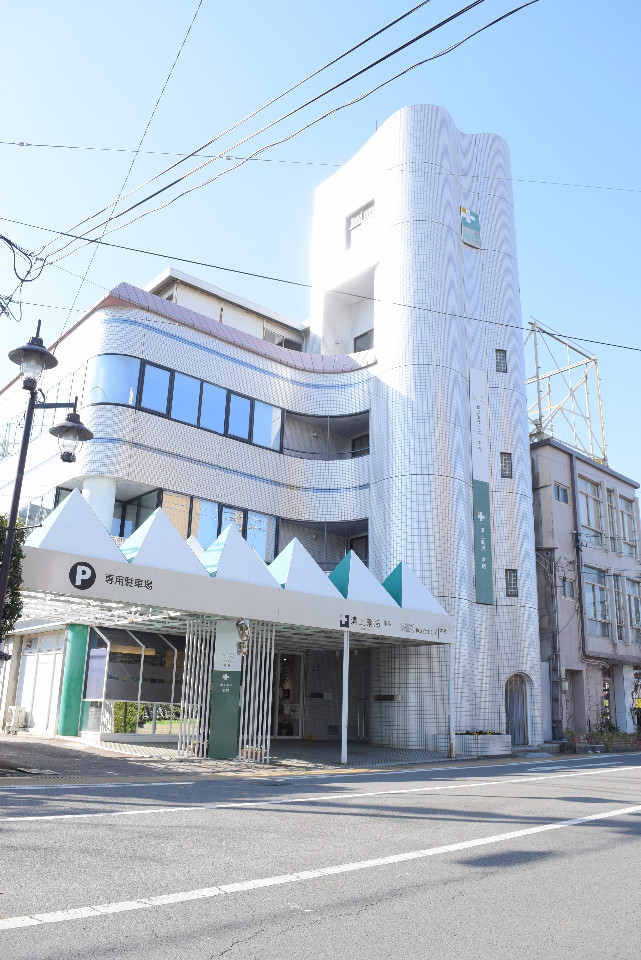 波打つような曲線を描く白いタイル張りの外壁が特徴的な多層階のビルで、1階部分には三角形が連なるユニークな屋根の駐車場の入り口があり、壁面には緑色の看板が設置されている建物の外観写真