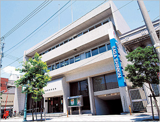 白いタイル貼りの多階建てビルの前に、「SAGA SHINKIN」と白抜き文字で書かれた鮮やかな水色の縦長看板が設置されており、街路樹の緑がビルに彩りを添えている佐賀信用金庫の店舗外観を斜め下から見上げた写真