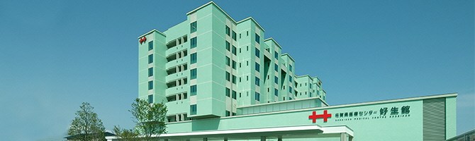 淡いグリーンの外壁が特徴的な巨大な病院建築が建ち並び、手前の建物には赤い十字のロゴマークとともに「佐賀県医療センター 好生館」という名称が大きく記された、施設の全景を捉えた写真