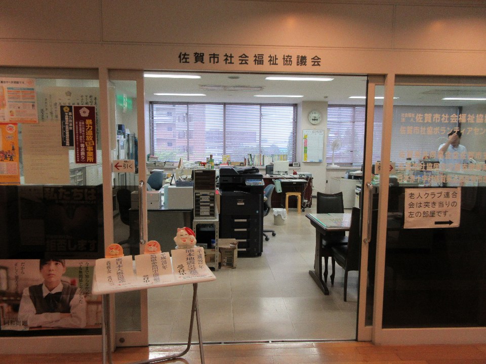 「佐賀市社会福祉協議会」の看板が掲げられた入口越しに、コピー機や事務机が並ぶ執務室が見え、手前には義援金受付の案内やポスターが掲示された、窓口付近の写真
