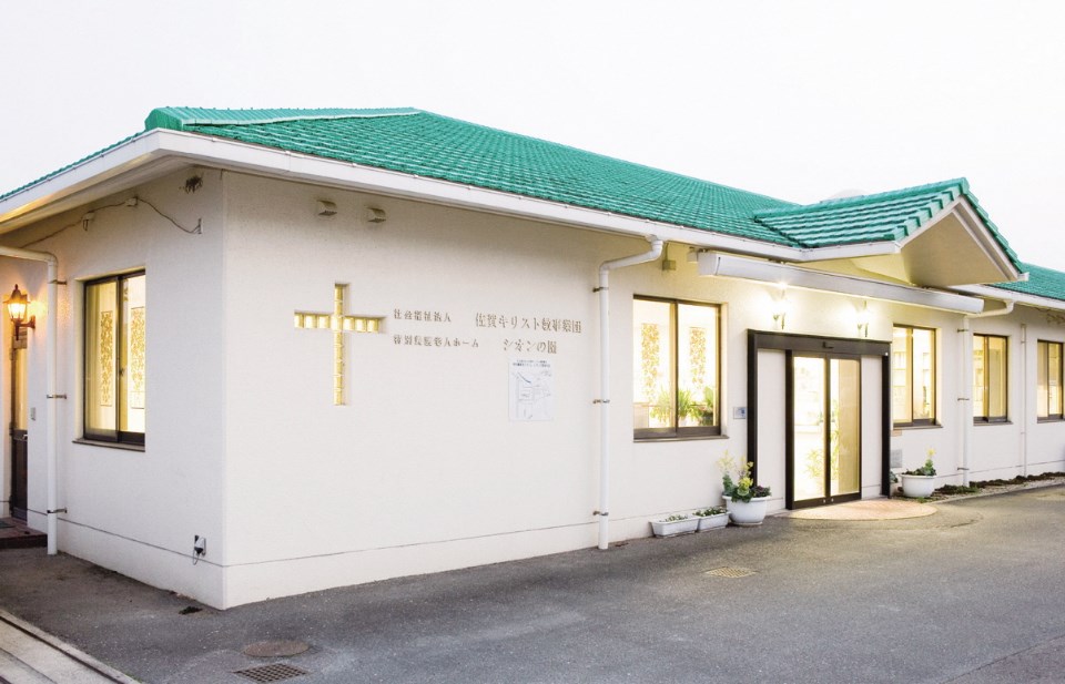 緑色の屋根と白い壁が特徴的な平屋建ての建物外観で、壁面には十字架の装飾と社会福祉法人佐賀キリスト教奉仕団特別養護老人ホーム シオンの園という施設名が記された、清潔感のある静かな佇まいの写真