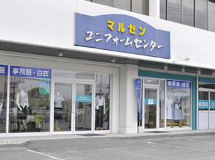 白い建物の壁面に「マルゼン ユニフォームセンター」と書かれた大きな青い看板が掲げられ、ガラス張りの店舗入口からは事務服や白衣を着用したマネキンが並んでいる様子が見えるマルゼンユニフォームセンターの外観写真