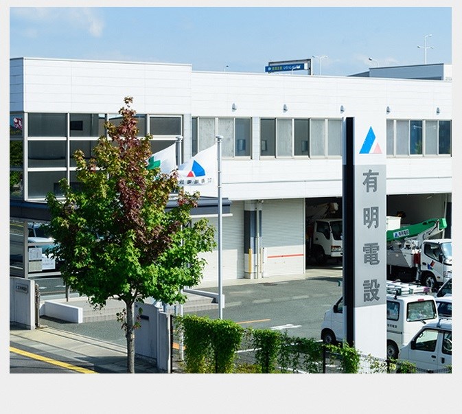 青と赤の三角形を組み合わせたロゴマークと「有明電設」の社名が記された高い自立看板が手前に立ち、奥には社旗が掲げられた白い二階建ての社屋と、高所作業車などの車両が並ぶ駐車場が広がっている会社の外観写真
