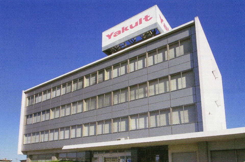 屋上に赤文字で「Yakult」と大きく書かれた白い看板を掲げ、規則正しく窓が並ぶグレーの外壁が特徴的な4階建ての自社ビルを、地上から見上げるような角度で捉えた写真