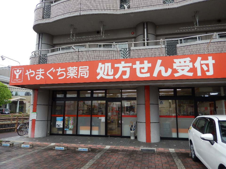 グレーのタイル貼りのマンション1階に位置し、鮮やかなオレンジ色の看板に白抜き文字で「やまぐち薬局 処方せん受付」と大きく表示されており、全面ガラス張りの店舗前にはレンガ調の駐車場が広がる調剤薬局の外観の写真