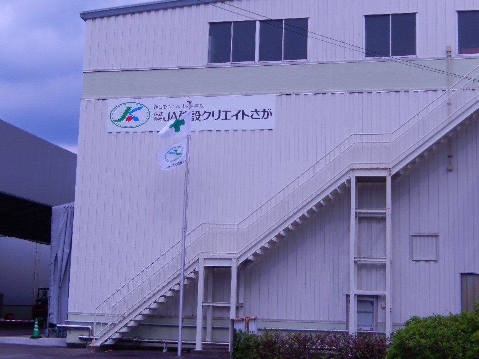 薄いベージュのトタン壁が印象的な大きな倉庫のような建物に、緑色のロゴマークと「株式会社JA建設クリエイトさが」という社名が書かれた看板が掲示されたクリエイト佐賀の外観写真