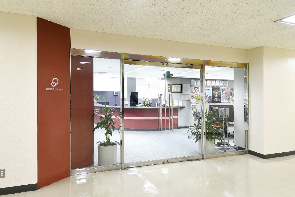 左側の壁面に円を重ねたロゴと「佐賀広告センター」の文字が白く記され、ガラス張りの自動ドア越しに曲線を描く受付カウンターや室内の観葉植物が見える、明るく清潔感のあるオフィスエントランスを捉えた写真
