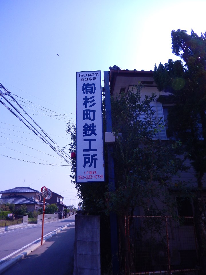 住宅街の道路脇に立つ「有限会社杉町鉄工所」と記載された白地の大きな看板を写した写真