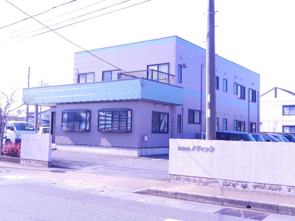 モダンな2階建て社屋で、手前には「株式会社メディック」と刻まれた白い石造りの門柱と駐車場が広がり、整然とした佇まいを見せる事務所の建物写真