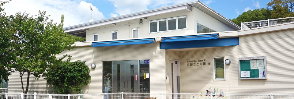 青いオーニングがせり出した白い横縞の外壁を持つ平屋建ての建物に、社会福祉法人旭福祉会「日新こども園」の文字が刻まれ、ガラス張りの正面玄関の横には掲示板や植栽、白いフェンスが設置されている、晴天の下の施設外観写真