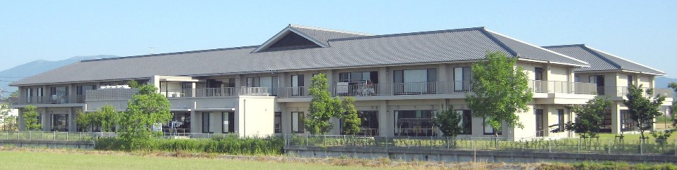 青空の下、和風屋根を持つ二階建ての白壁建物が草地と若木の並木越しに見え、背景に山並みが広がる特別養護老人ホーム春庵の外観写真