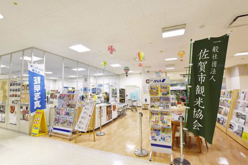 「一般社団法人佐賀市観光協会」と書かれた深緑色ののぼり旗が手前に立ち、明るく開放的な観光案内所の窓口付近を捉えた写真
