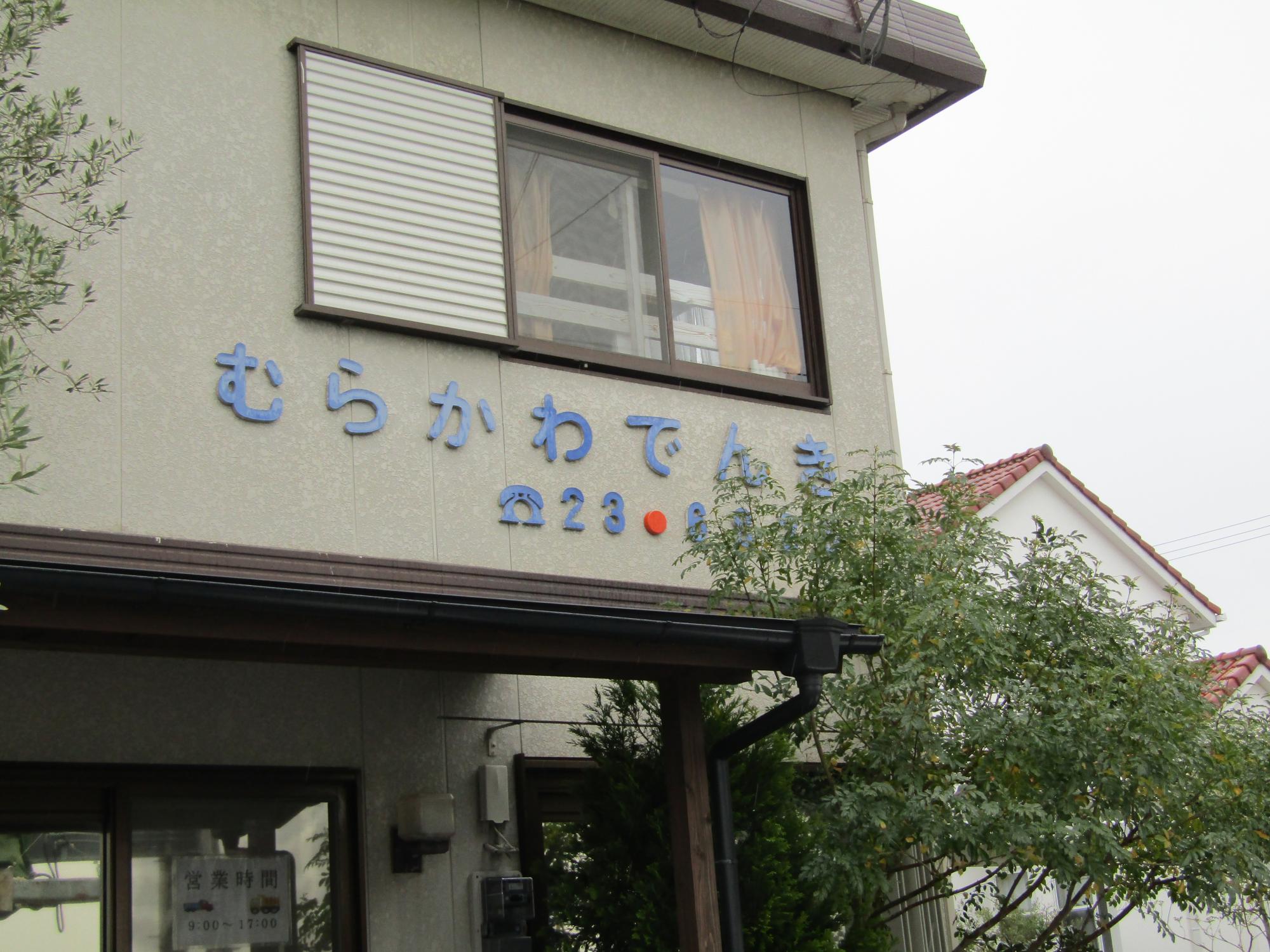 落ち着いたベージュ色の外壁を持つ2階建て住宅兼店舗の壁面に、丸みを帯びた青い文字で「むらかわでんき」という店名と電話番号が掲げられた村川電機の外観写真