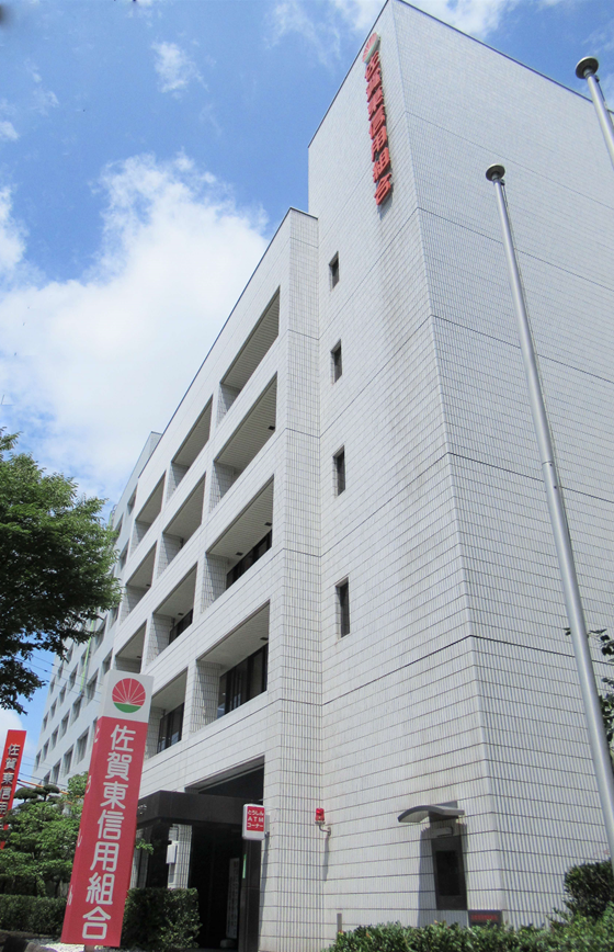 白いタイル貼りの大きなビルの壁面に赤い文字で「佐賀東信用組合」という看板が掲げられ、手前にも同じロゴと名称が入った赤い縦長看板が設置されている、佐賀東信用組合の外観写真