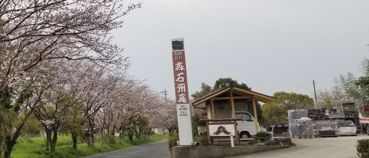 茶色い背景に白抜き文字で「有限会社 森石州瓦」と書かれた縦長の大きな社名看板が立っており、その奥には瓦屋根の小さな建物や資材が積まれた作業場の写真