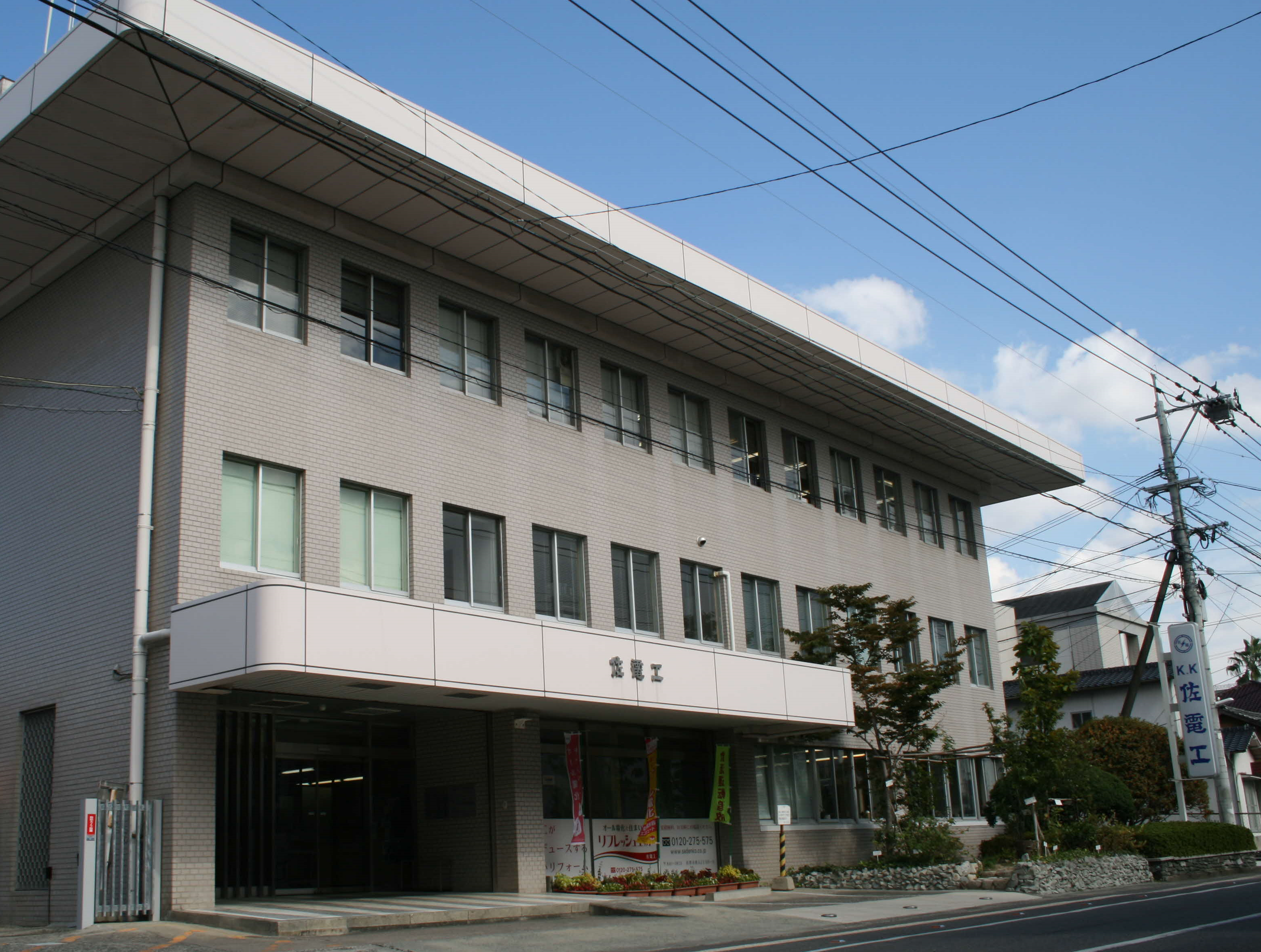 薄いグレーのレンガ調の壁面に、突き出した白い庇の中央に「佐電工」という文字が掲げられた3階建ての大きなビルの佐電工外観写真