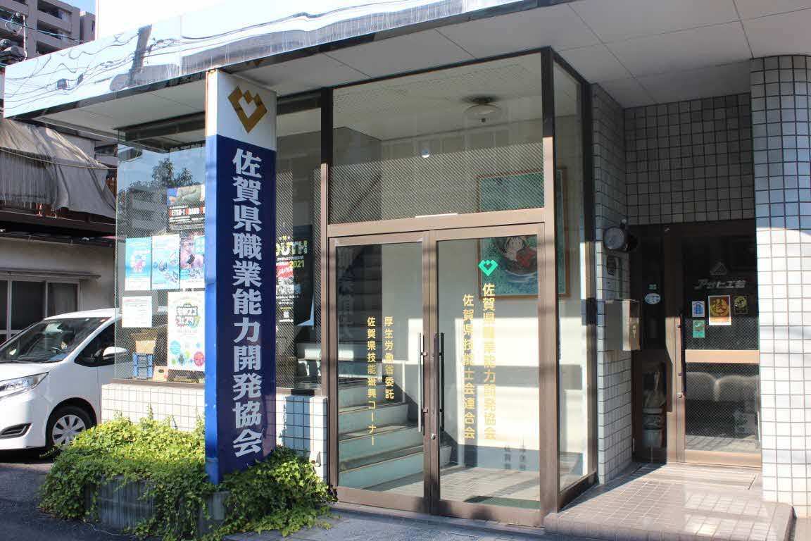 建物の入り口に「佐賀県職業能力開発協会」と大きく書かれた青い看板が立ち、ガラス張りのドアには厚生労働省委託の技能振興コーナーなどの案内が記された、佐賀県職業能力開発協会の外観写真