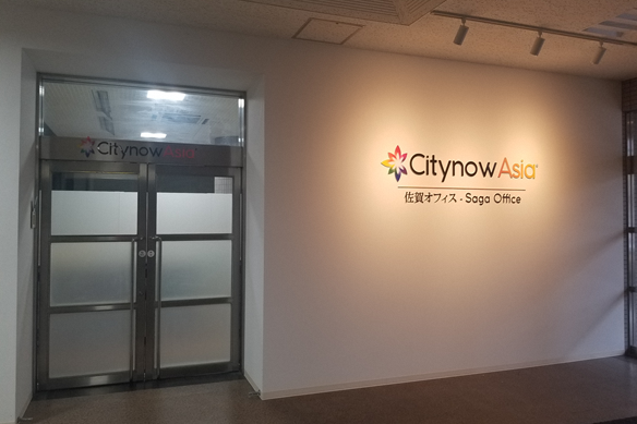 カラフルなロゴマークと「Citynow Asia 佐賀オフィス - Saga Office」という文字がライティングで浮かび上がり、明るいベージュの壁面の写真
