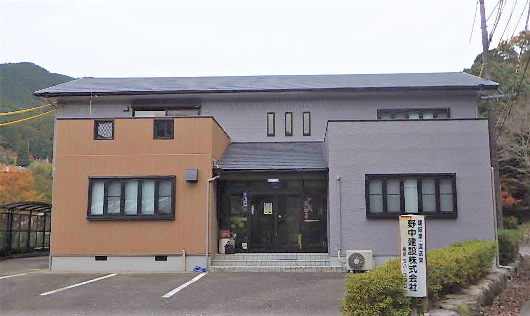 落ち着いたグレーとベージュの2トーンカラーが印象的な2階建てのオフィスビルで、正面には「建設業・運送業 野中建設株式会社」と記された縦長の白い看板が立ち、山あいに建つ野中建設社屋の外観写真