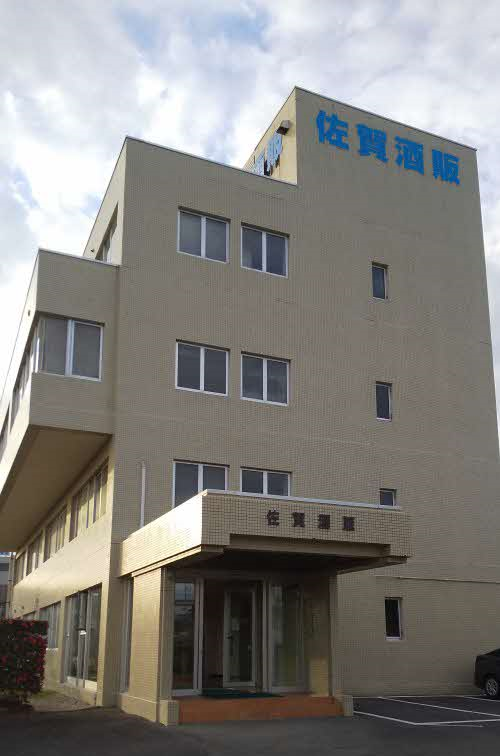 薄いベージュ色のタイルの外壁に、青い文字で「佐賀酒販」と大きく書かれた看板が最上部に見え、4階建て佐賀酒類販売株式会社のビルを斜め前方から捉えた写真