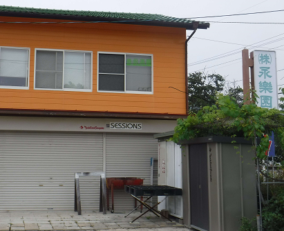 鮮やかなオレンジ色の外壁が目を引く2階建ての建物で、1階の閉まったシャッターの上には「SESSIONS」というロゴが記されており、建物の右側には「株式会社永楽園」と書かれた縦長の白い看板が木々に囲まれて立っている写真