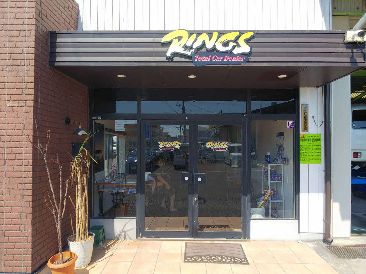 茶色のレンガ壁と白い外装が組み合わされた建物の入り口に、黄色い筆文字風の「RINGS」というロゴと赤字の「Total Car Dealer」が書かれた黒い看板が掲げられたリングス株式会社の外観写真