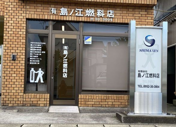 茶色のレンガ調の壁面に「有限会社島ノ江燃料店」の切り文字看板と、ロゴマークや電話番号が記された自立式のシルバーの看板が設置された、ガラス扉と大きな窓がある店舗の正面入り口を撮影した写真