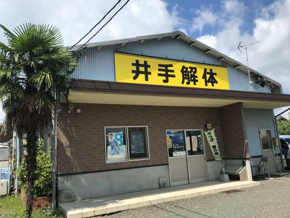 青空の下に立つ「井手解体」と書かれた大きな黄色い看板が掲げられた平屋の建物と、その左側に植えられた背の高いヤシの木、そして建物の窓や入り口付近に貼られたポスターやのぼり旗が映り込んでいる写真