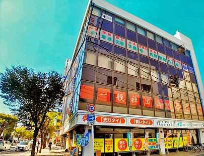 大きな窓が特徴的な多層階ビルの外観で、1階部分に「売りタイ」「買いタイ」というオレンジ色の目立つ看板や不動産仲介の黄色いのぼりが並んだ株式会社ソロンの外観写真