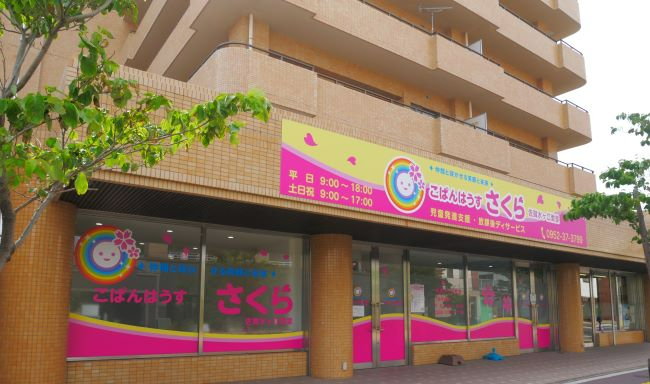 茶色のタイル貼りのマンション1階部分に入居する施設で、黄色とピンクの鮮やかな看板に「こぱんはうす さくら」という施設名と笑顔の虹色アイコンが描かれた、ホームページ用水ケ江教室の外観写真
