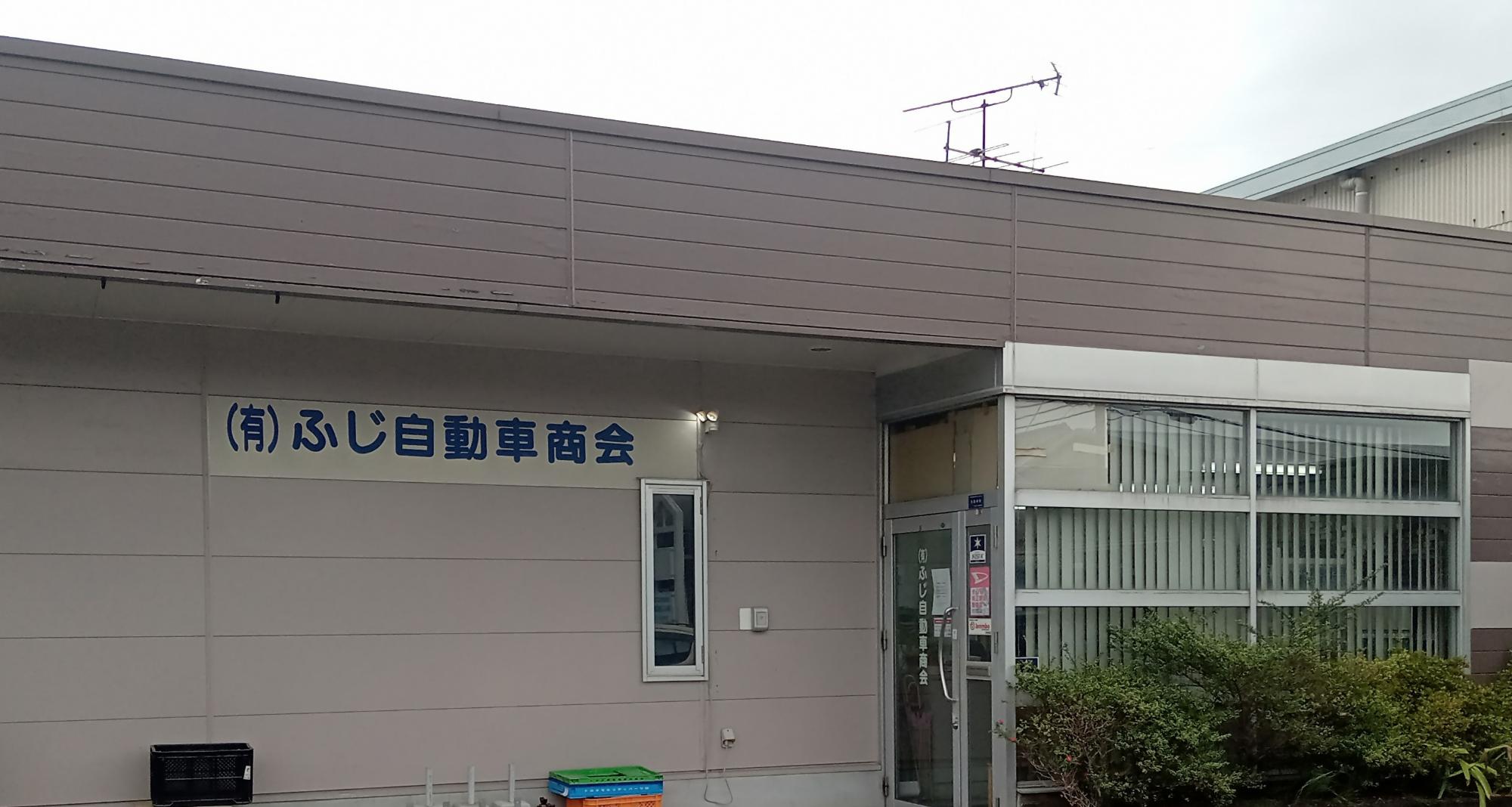 落ち着いたグレーの平屋建て事務所で、壁面には青い文字で「有限会社ふじ自動車商会」と社名が記され、ガラス張りの入口付近にはブラインドの付いた窓や低木が並ぶ、ふじ自動車商会の外観写真