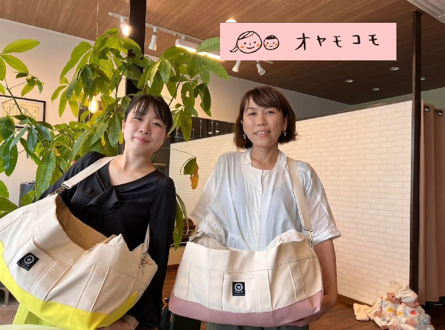 観葉植物が置かれた室内で、大きなマザーズバッグを肩にかけた二人の女性が笑顔で並んでおり、右上には親子のイラストと「オヤモコモ」というロゴ、背景の右下には赤ちゃんの人形が置かれている様子を捉えた写真