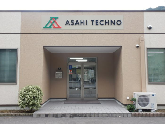 中央のガラス扉の入り口の横には低木やエアコンの室外機が設置されている、ベージュと白のツートンカラーの外壁を持つ建物の正面に、緑と赤を組み合わせた「A」のロゴと「ASAHI TECHNO」の黒い文字が掲げられた建物外観の写真