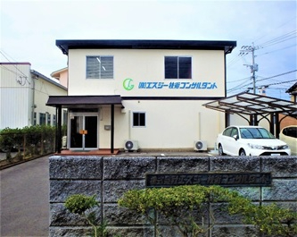 薄いベージュ色の外壁に「株式会社エスジー技術コンサルタント」の社名とロゴが青色で記された2階建ての社屋と、その手前に積まれた石造りの門壁、右側のカーポートに停車している白い乗用車を、穏やかな曇り空の下で捉えた写真
