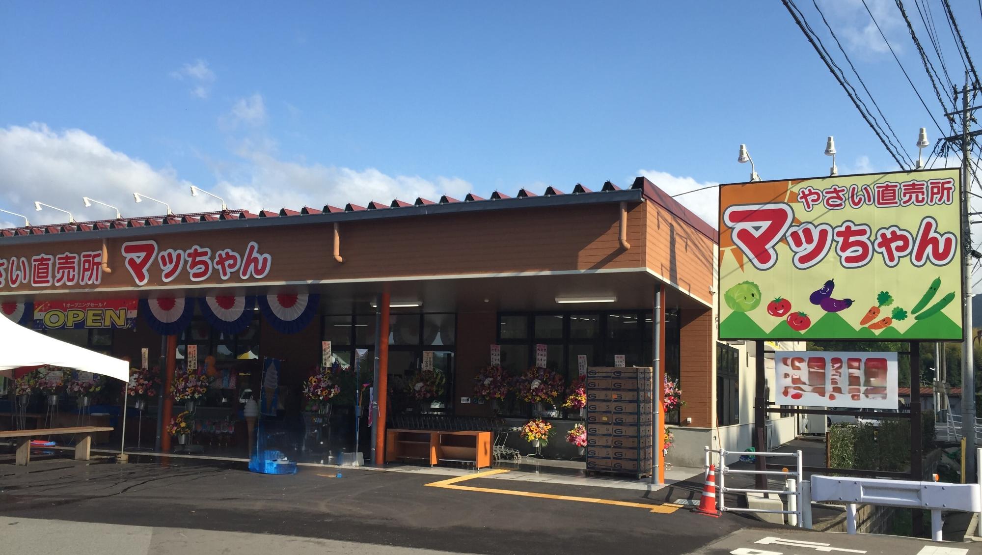 茶色の外壁の店舗前に「やさい直売所 マッちゃん」と大きく書かれた黄色い看板が立ち、店頭には開店祝いの華やかな花輪が多数並んでいる、マッちゃん店舗の外観写真
