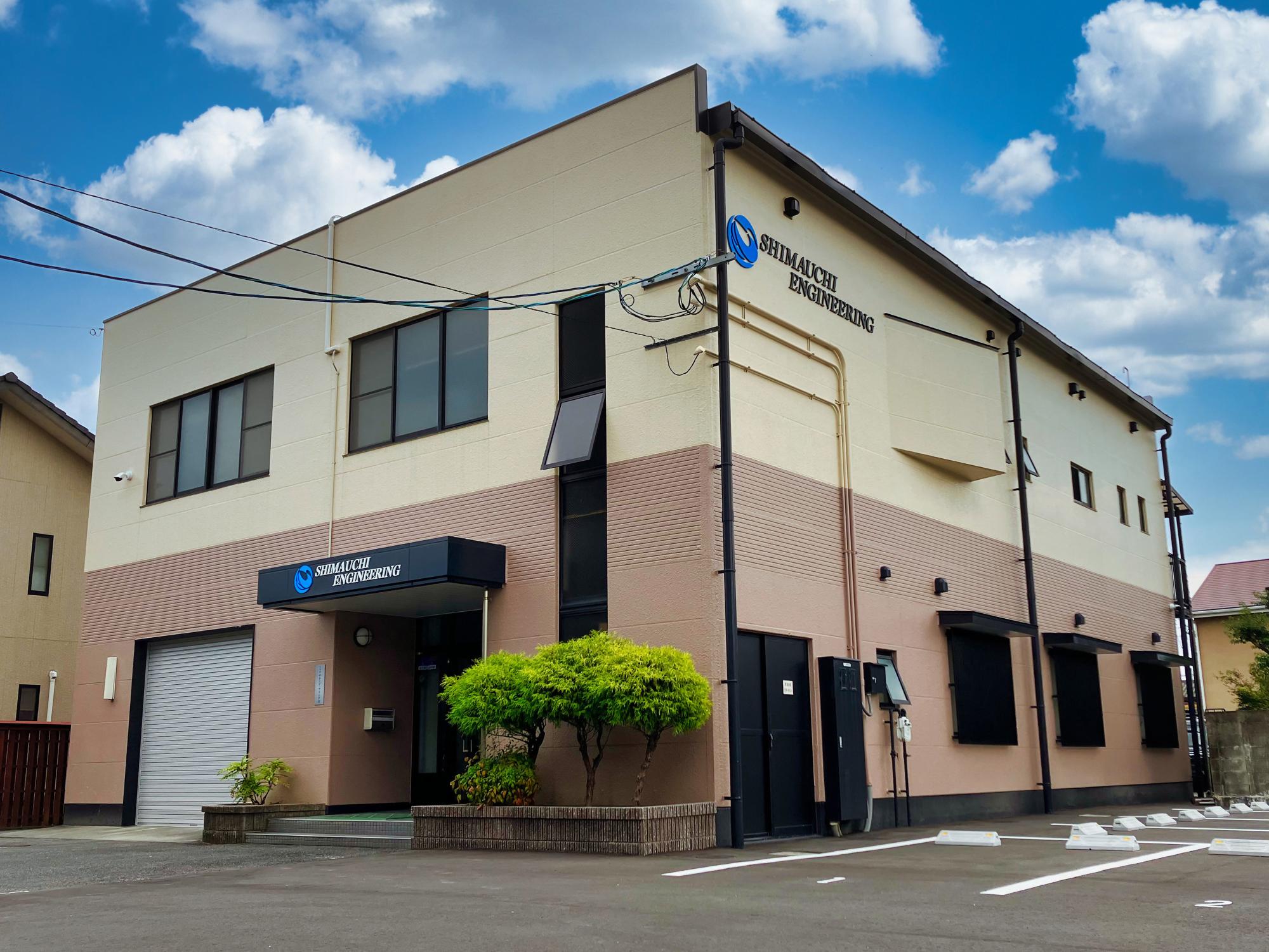 ベージュと茶色のツートンカラーの壁面が特徴的な2階建てのオフィスビルで、入り口の庇や壁面に「SHIMAUCHI ENGINEERING」という青いロゴ入りの会社名が掲げられている様子を斜め前から捉えた写真