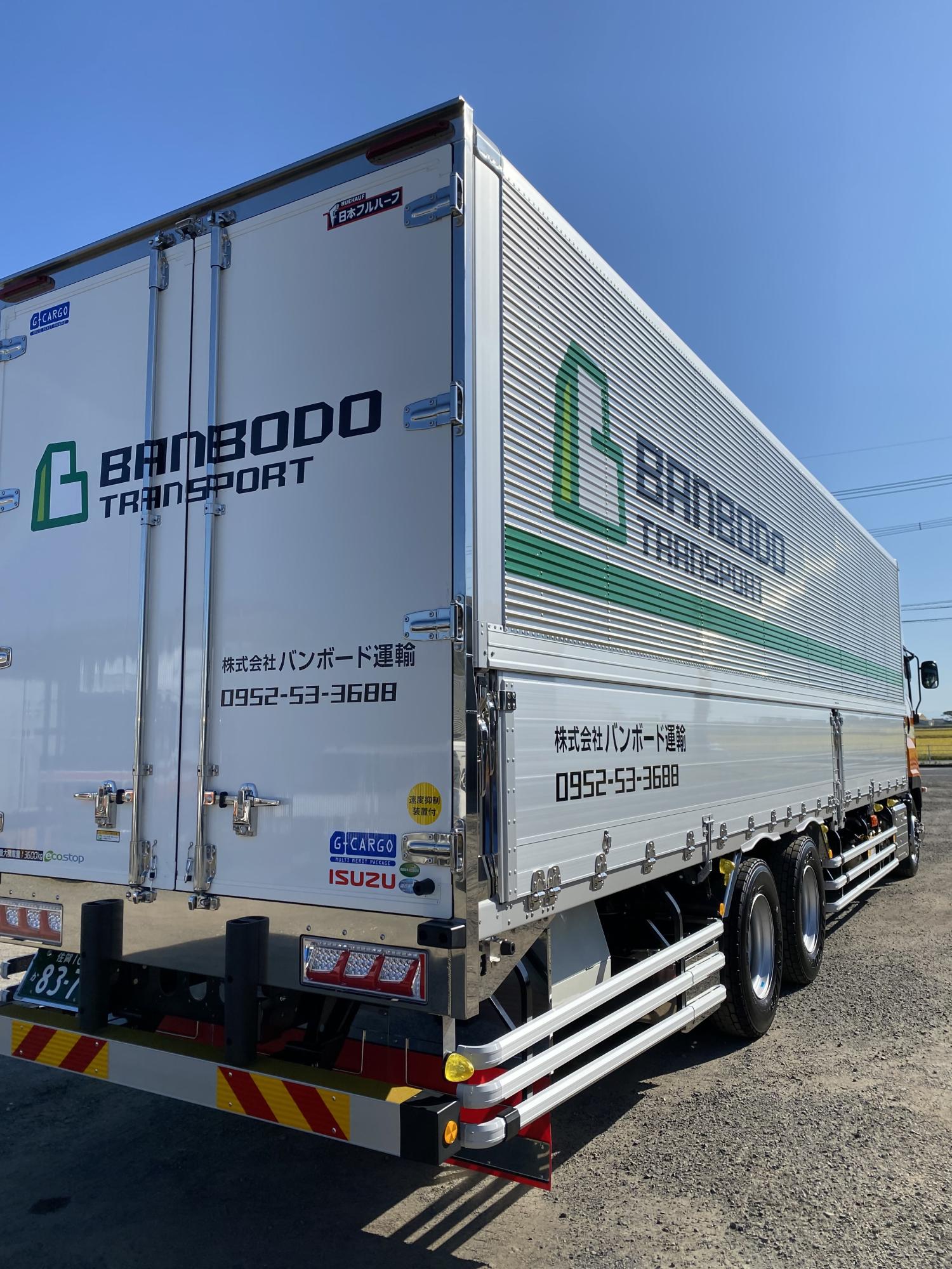 白く輝く大型のアルミウィングトラックで、車体側面と背面には「BANBODO TRANSPORT」という英字ロゴや「株式会社バンボード運輸」の社名、電話番号が黒と緑色で大きく記された車両の写真