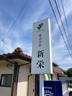 カラフルなロゴマークと「SHIN-EI」、「株式会社 新栄」という社名が縦書きで記された看板を写した写真